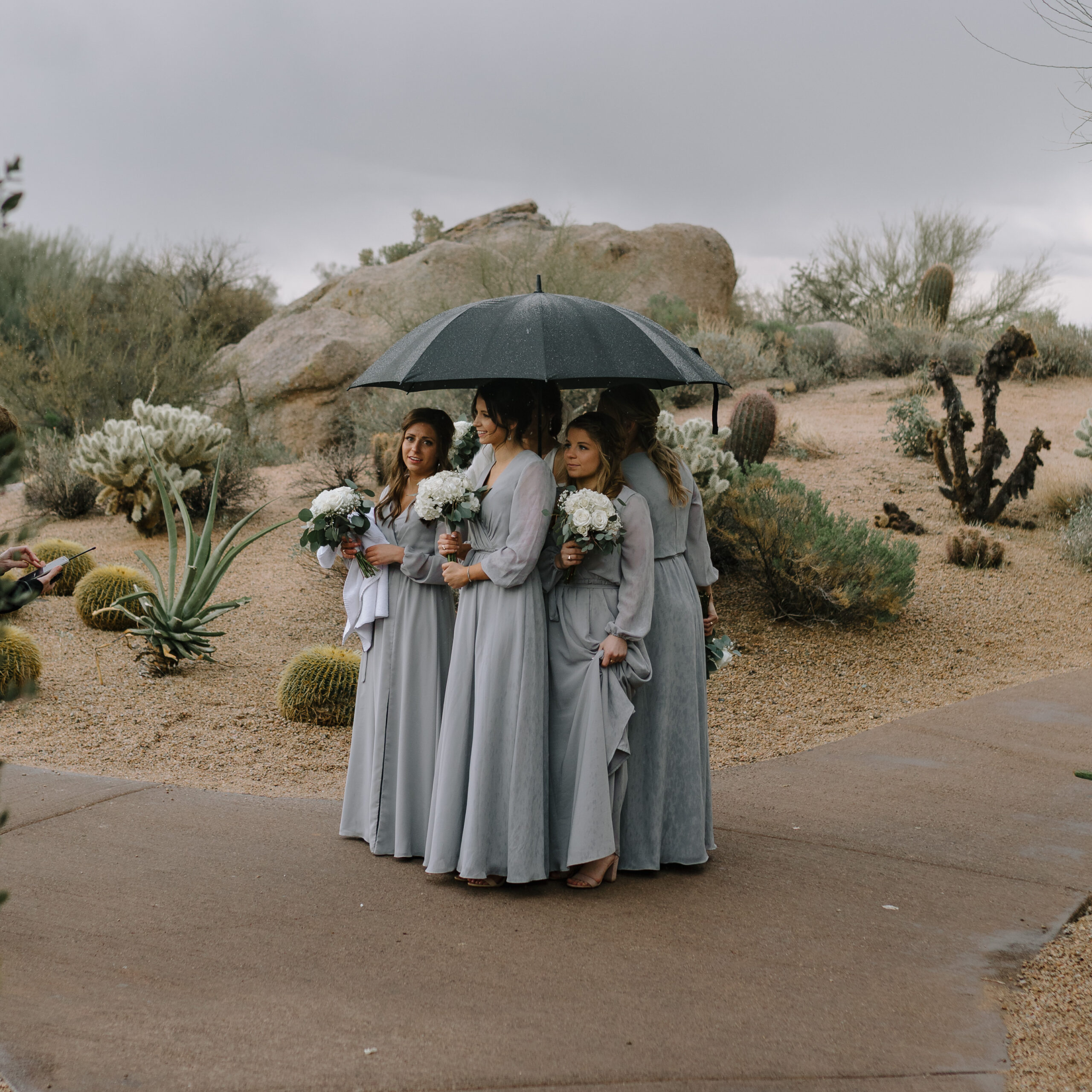 Rainy wedding day with bridesmaids sharing an umbrella among desert cacti a beautifully imperfect candid moment. How to relax in front of the camera (for wedding photos)