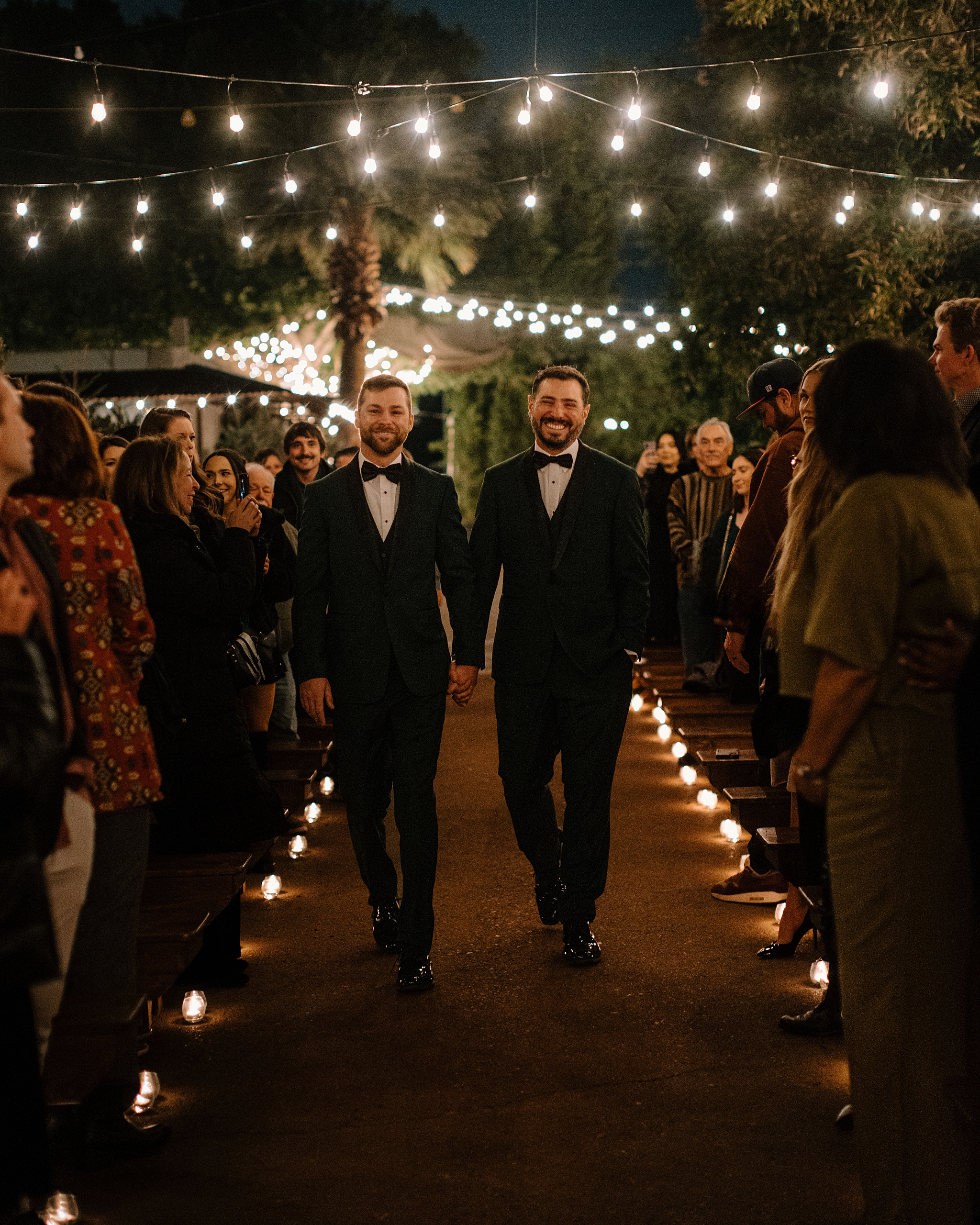 Two grooms walking into their outdoor candlelit wedding ceremony at Whitfill Nursery in Phoenix